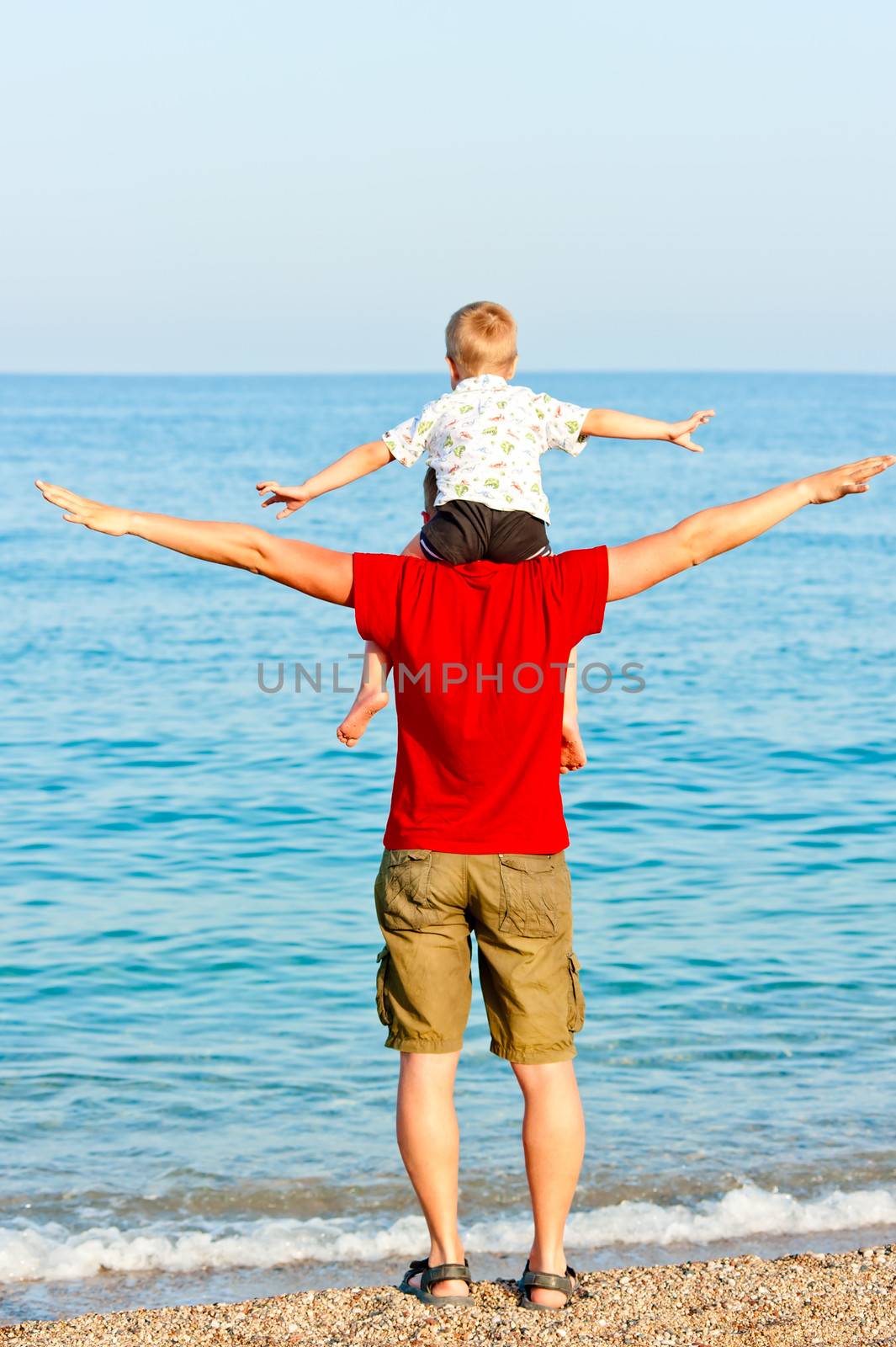 Son sits on his father's shoulders his arms to the side against the sea