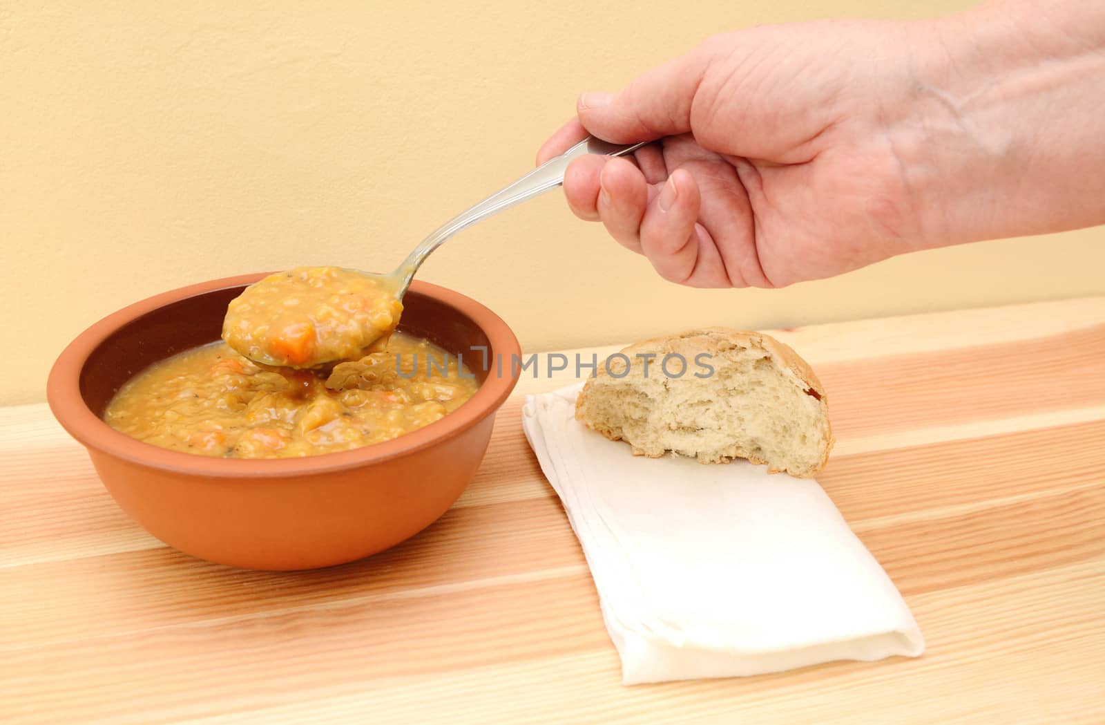 Hand lifts spoonful of vegetable soup from bowl with bread roll and napkin on table