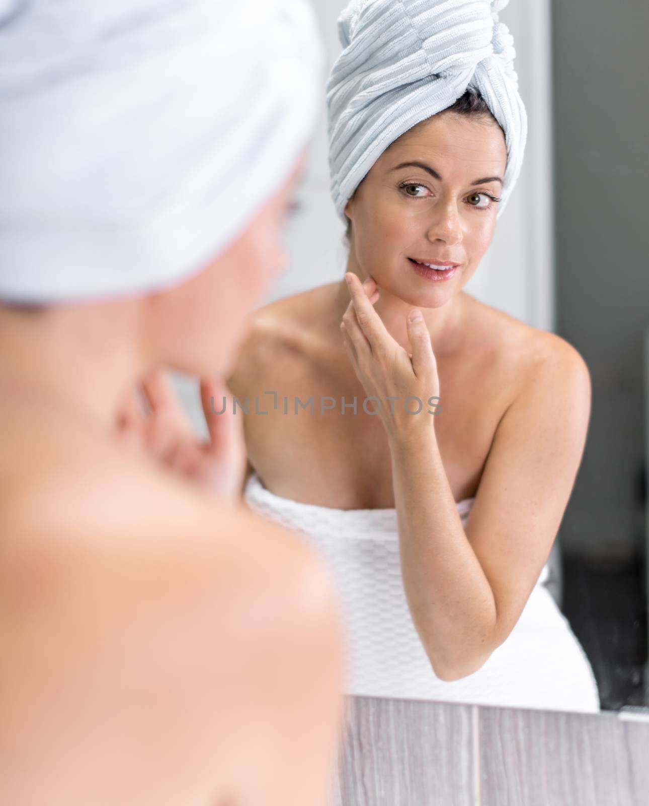 portrait of beautiful mature lady with a towel looking at her skin in the mirror