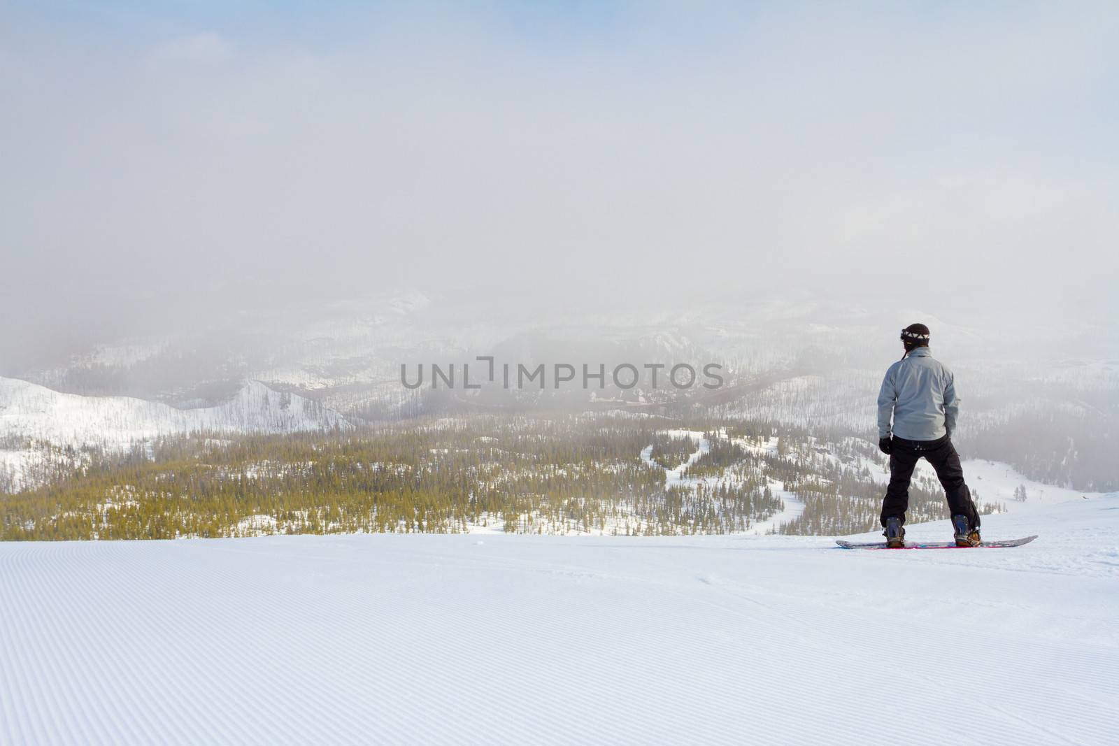 A man on a snowboard waits to drop in from the summit of a high elevation mountain.