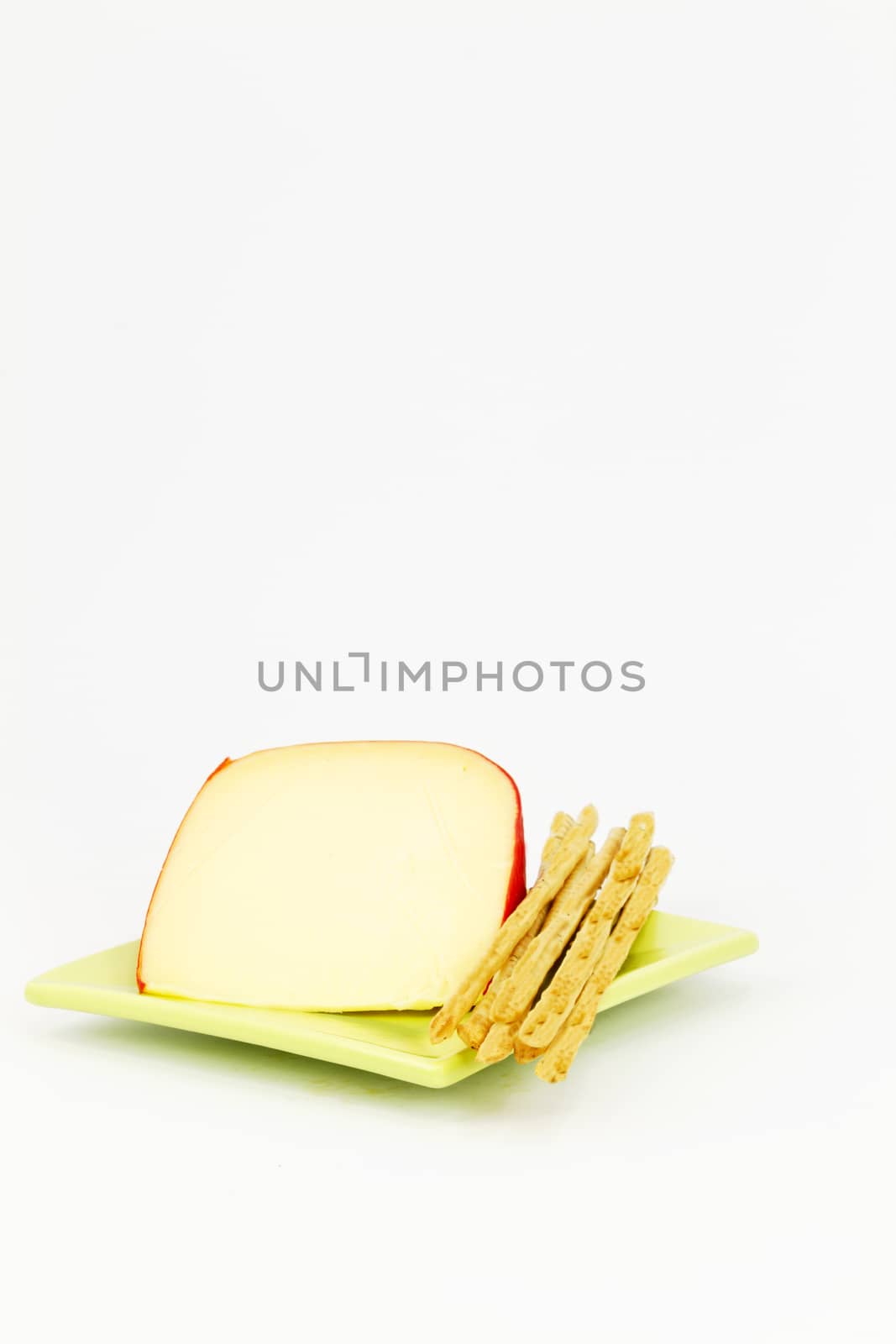 Slice of yellow cheese with red rind placed with breadsticks on green, square plate against white background; vertical image with large areas of copy space.