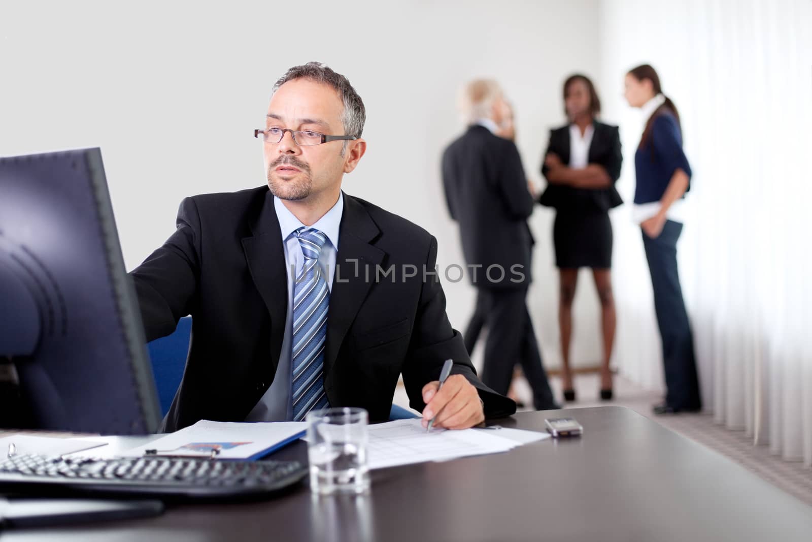 Handsome mature business man writing notes while using computer at office with colleagues