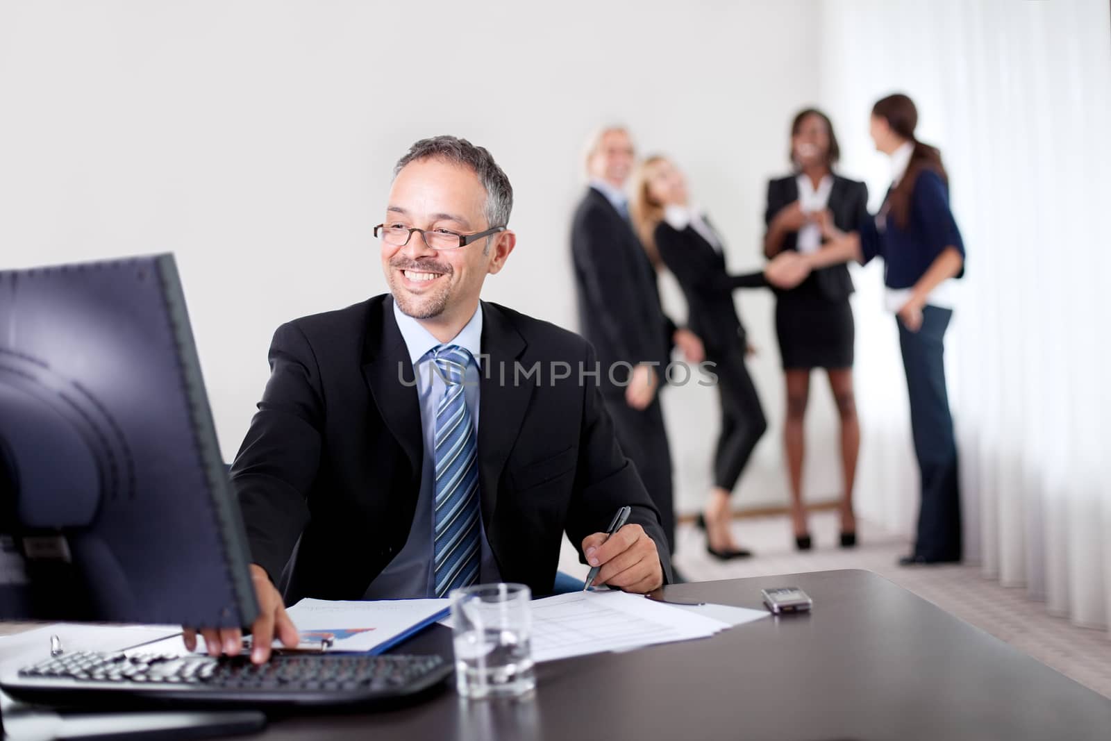 Confident happy businessman writing notes while using computer at office with colleagues in the background