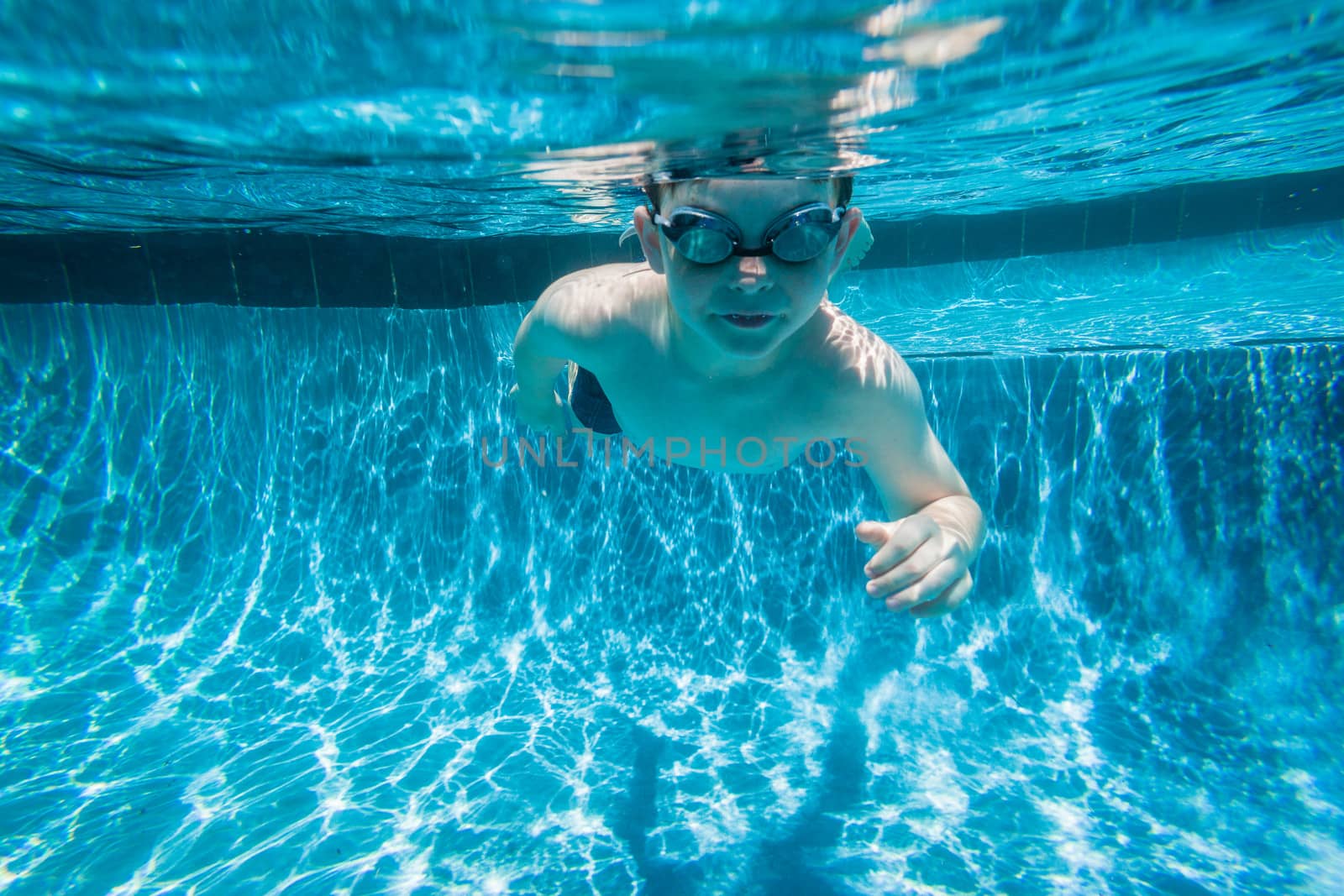 Young boy wearing goggles underwater in swimming pool.