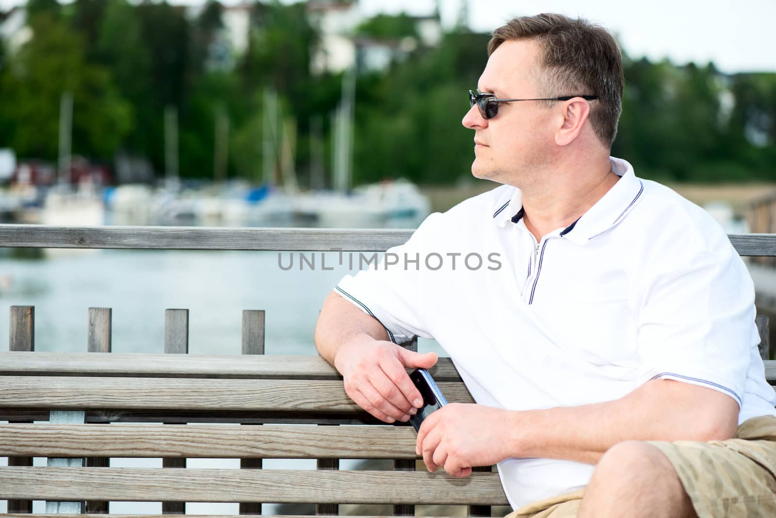 Mature man looking away and sitting on wooden bench with smart phone in hands