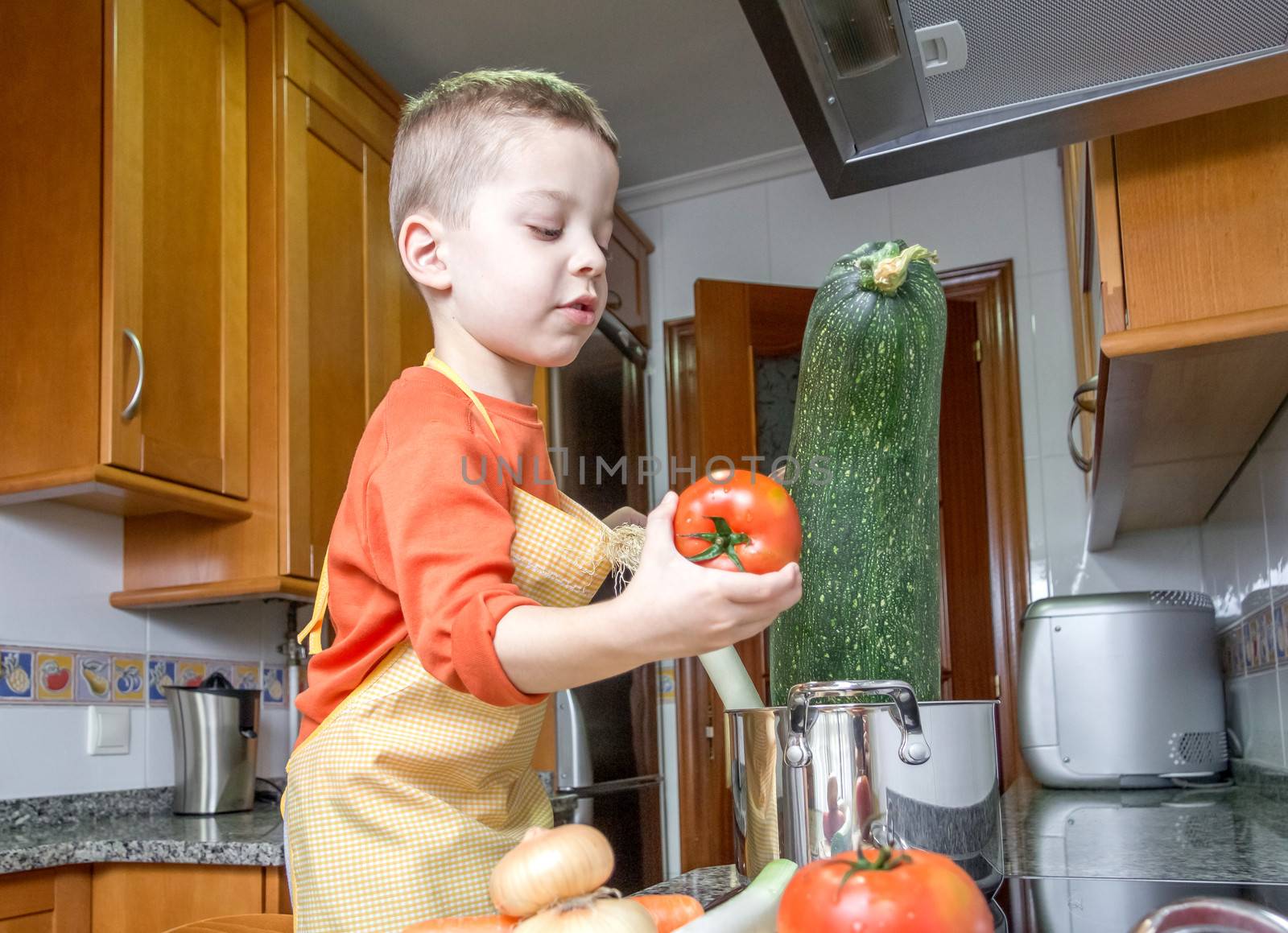 Cute child chef with apron cooking big zucchini and other vegetables in a pot on the kitchen