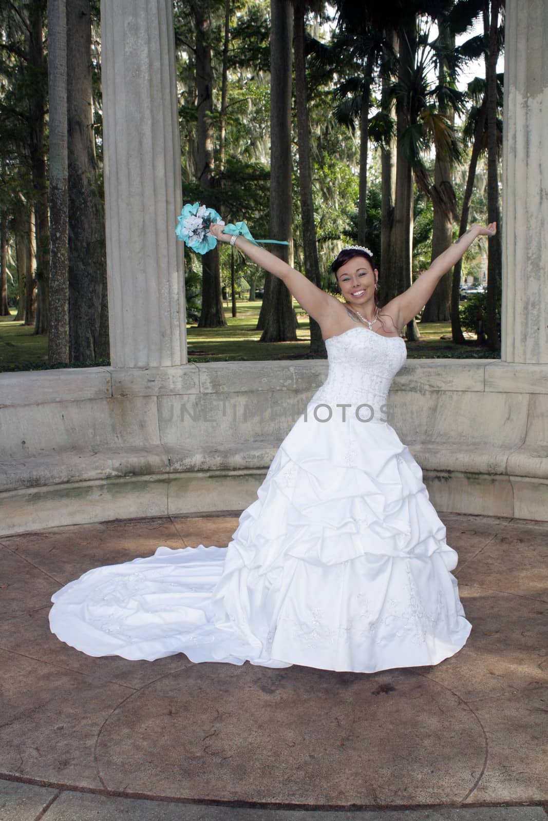 A lovely young bride outdoors wearing her wedding gown and holding her boquet, celebrates with her arms raised.