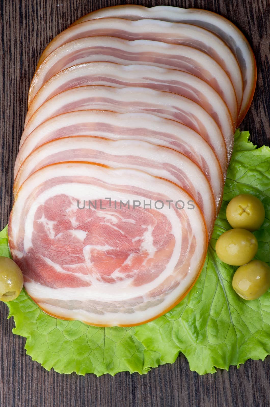 Slices of Delicious Smoked Ham Roll with Lettuce and Green Olives closeup on Wooden background