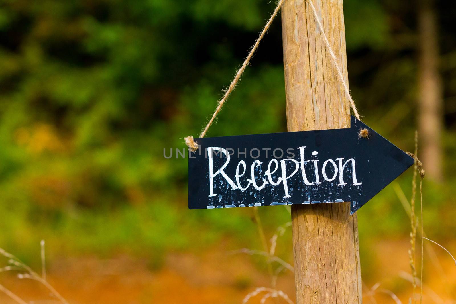 A wedding reception sign is made from a small chalkboard and has been outside in the rain. The sign is an arrow hung with twine.