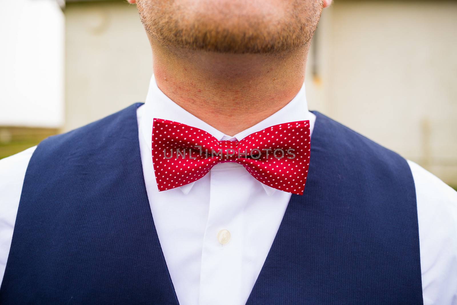 A fashionable groom wears a red and white bowtie with a navy blue vest on his wedding day.