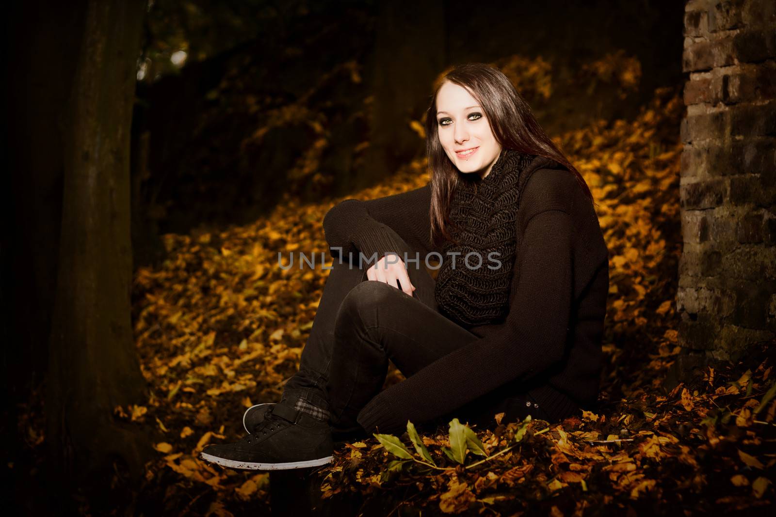 Beautiful woman in autumn leaves sitting outside in the darkness on the ground alongside a brick wall, with copyspace