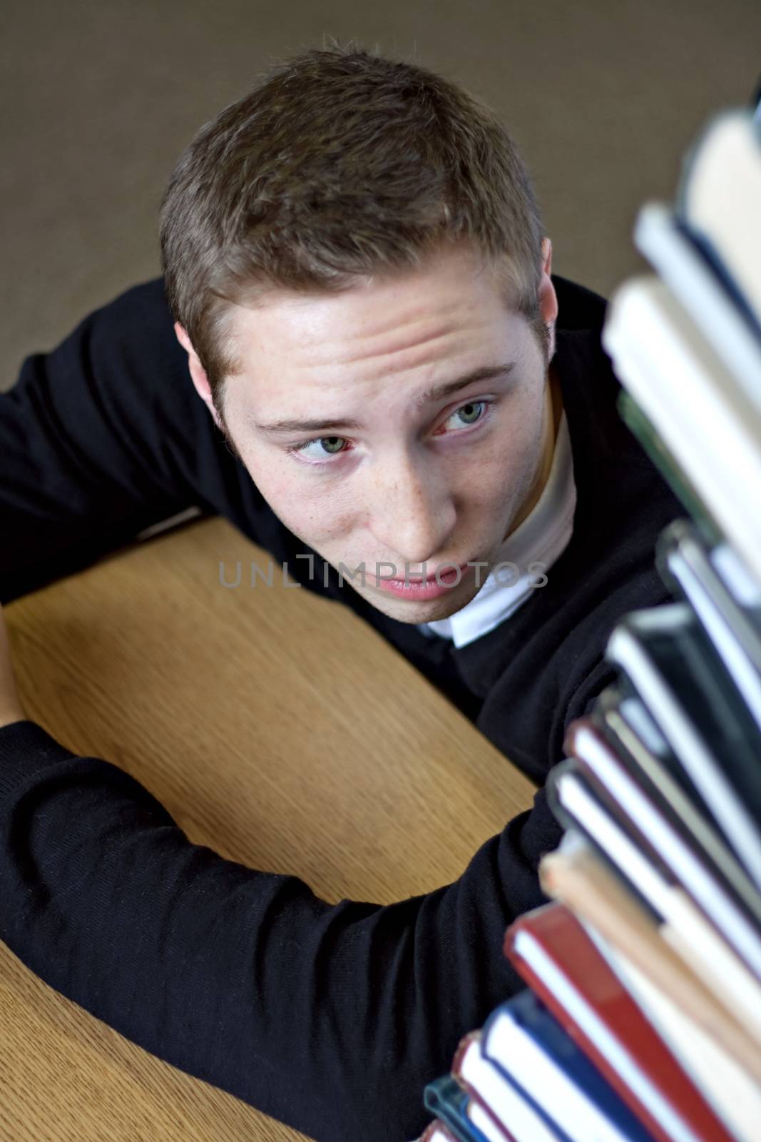 A frustrated student looks up at the high pile of textbooks he has to go through to do his homework assignment. Shallow depth of field.