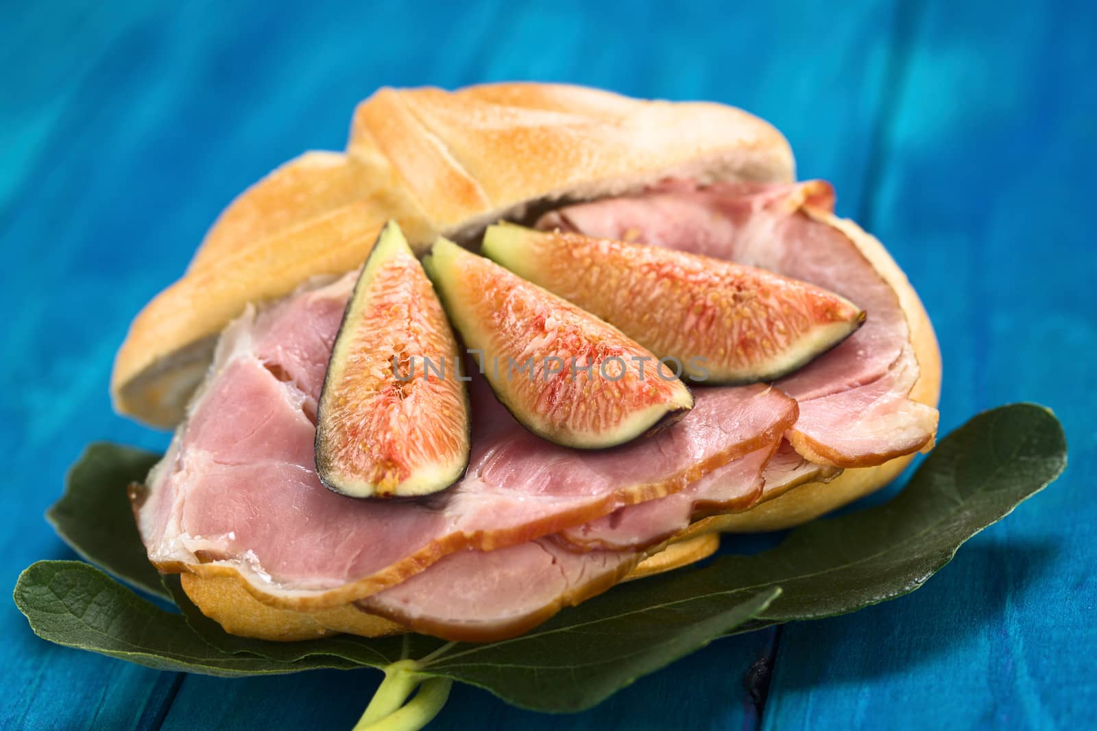 Fresh fig slices on smoked ham sandwich on fig leaves on blue wood (Selective Focus, Focus on the middle of the two fig slices on the left)