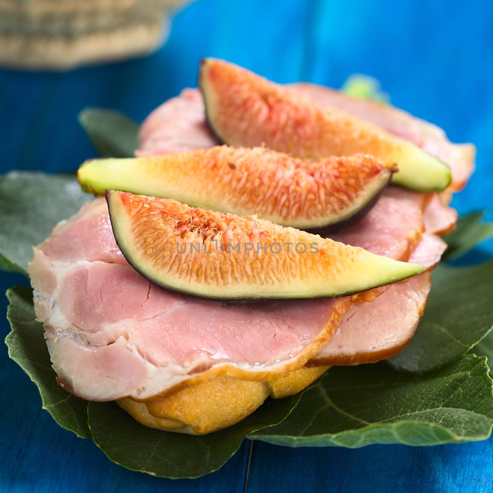 Fresh fig slices on smoked ham sandwich on fig leaves on blue wood (Selective Focus, Focus on the first fig slice)