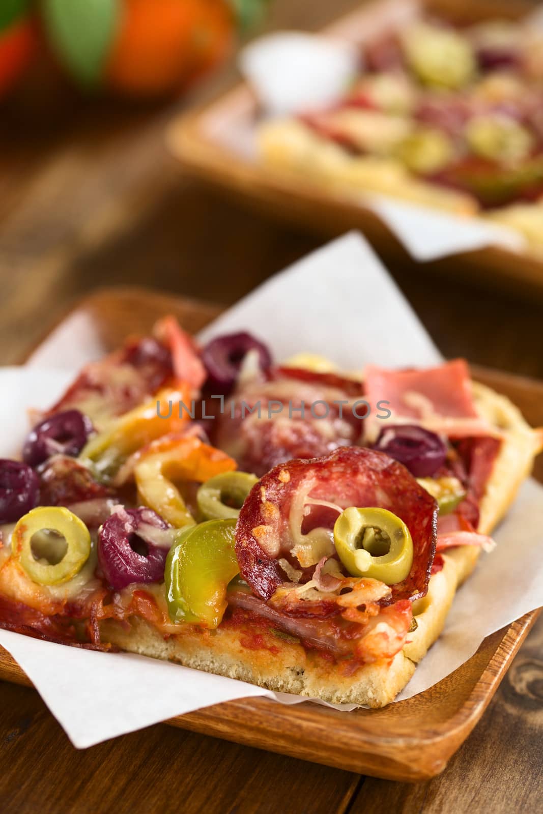 Fresh homemade pizza piece with tomato sauce, ham, salami, olives, bell pepper and cheese on top, served on sandwich paper on wooden plate (Selective Focus, Focus on the front of the green olive slice on the right)