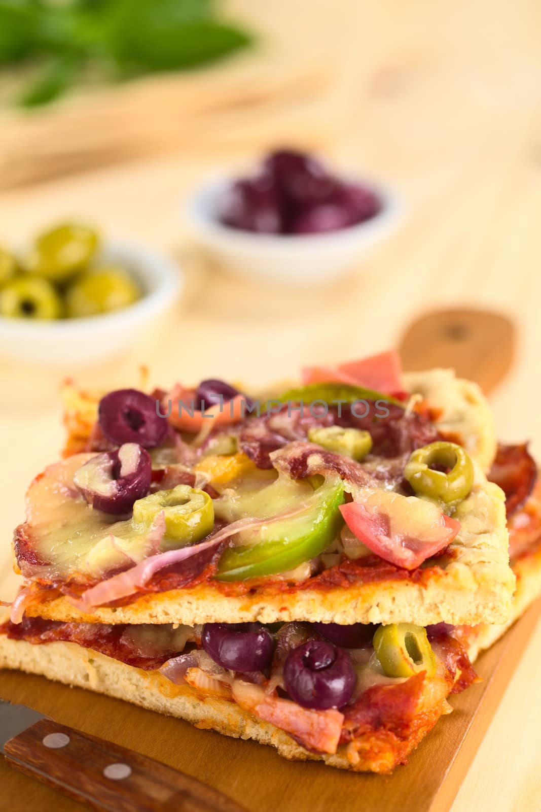 Fresh homemade pizza pieces with tomato sauce, ham, salami, olives, bell pepper and cheese on top on wooden board with knife (Selective Focus, Focus one third into the image)