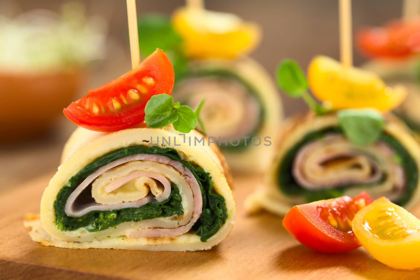 Crepe rolls as finger food filled with spinach and ham garnished with cherry tomato and watercress served on wooden board (Selective Focus, Focus on the upper part of the crepe roll) 