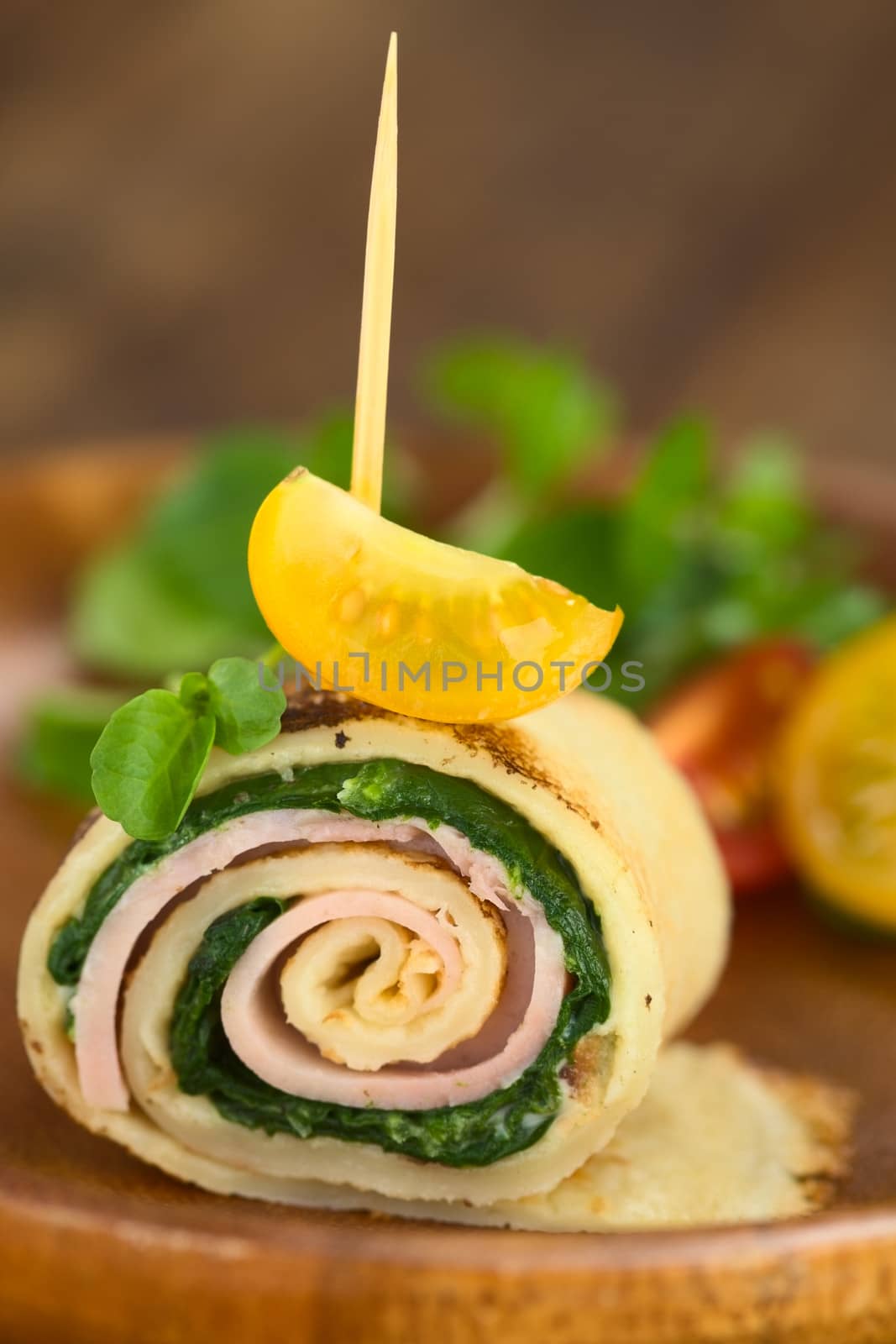 Crepe roll as finger food filled with spinach and ham garnished with cherry tomato and watercress served on wooden plate (Selective Focus, Focus on the upper part of the crepe roll and on the front of the cherry tomato on top) 