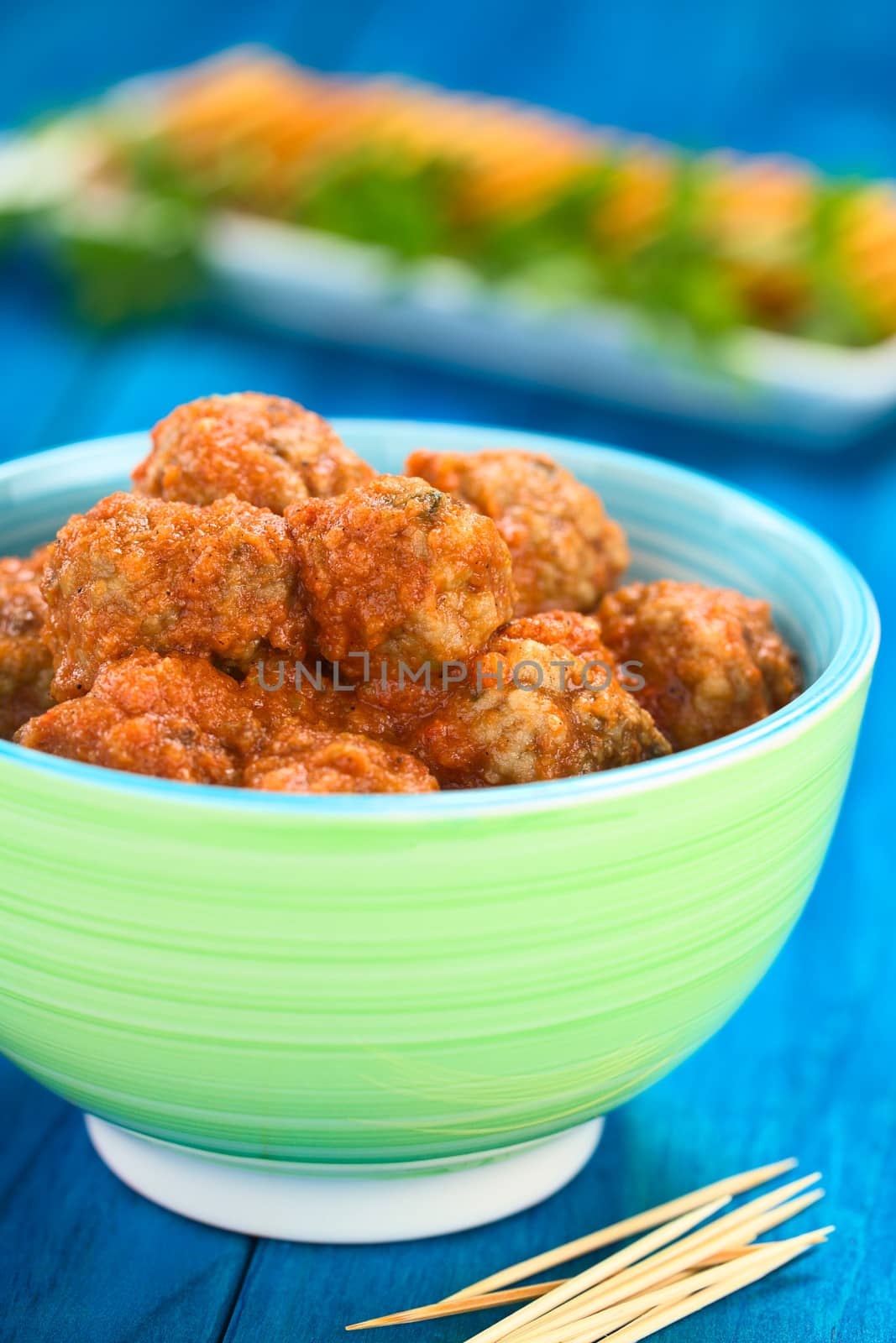 Spanish albondigas (meatballs) in tomato sauce in colorful bowl with toothpick in the front and crackers in the back on blue wood (Selective Focus, Focus on the meatballs on the top)