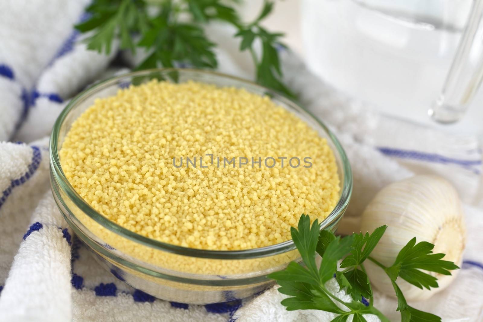 Raw couscous in glass bowl with garlic and parsley on the side, a jug of water in the back, on kitchen towel (Selective Focus, Focus one third into the couscous)