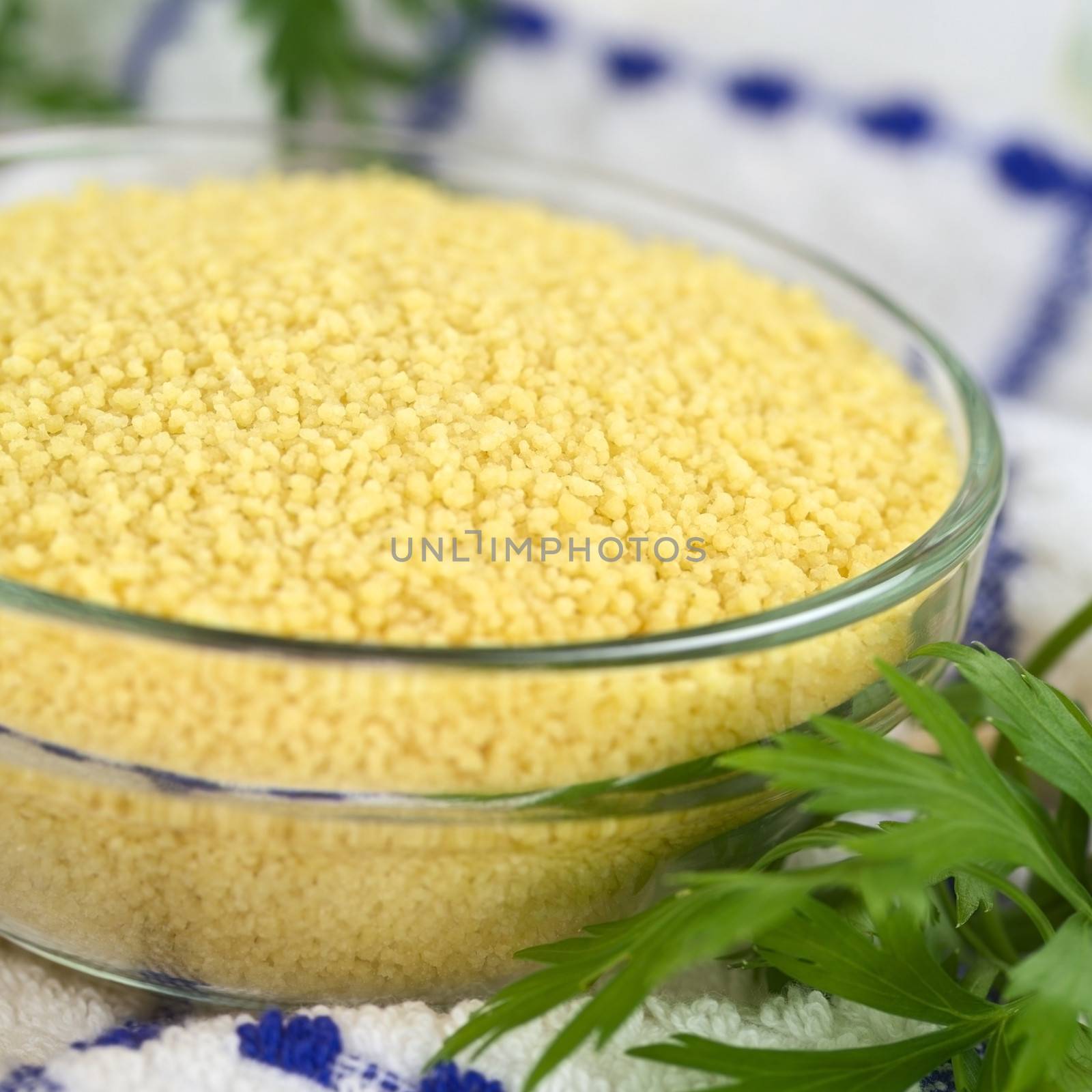 Raw couscous in glass bowl with parsley on the side on kitchen towel (Selective Focus, Focus one third into the couscous)