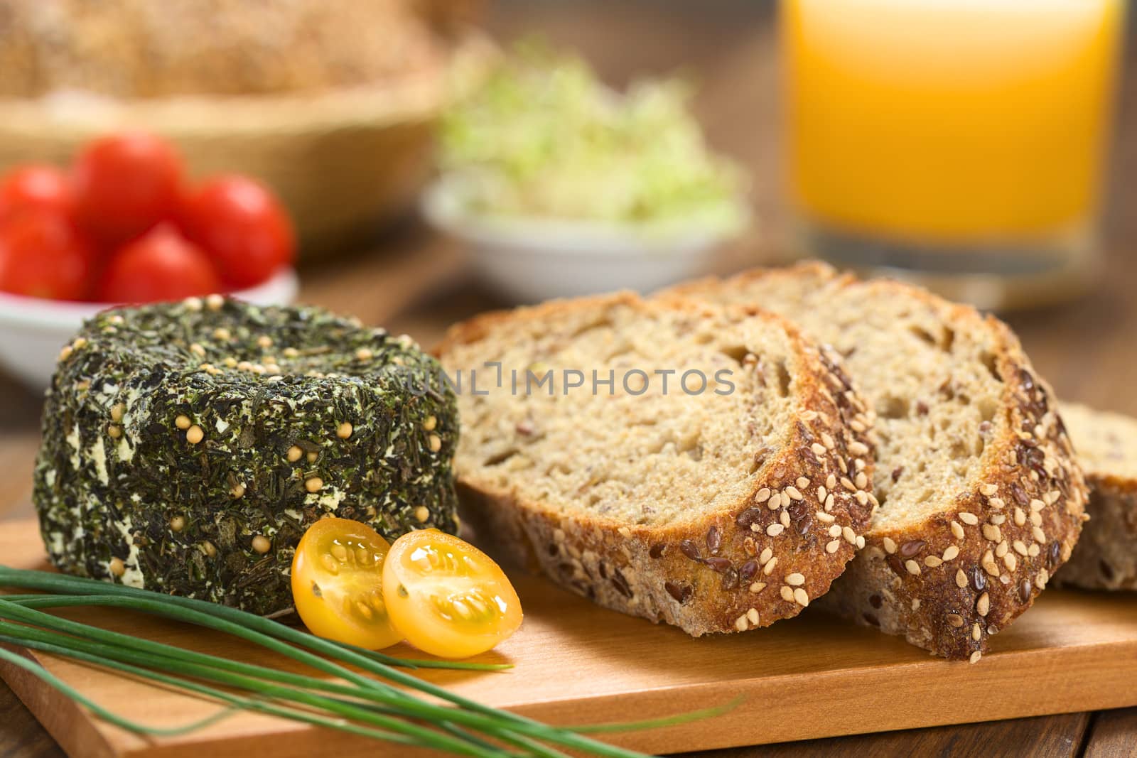 Goat cheese covered with herbs, chives, yellow cherry tomato and slices of wholegrain bread on wooden board (Selective Focus, Focus on the front of the cheese and the bread slices)