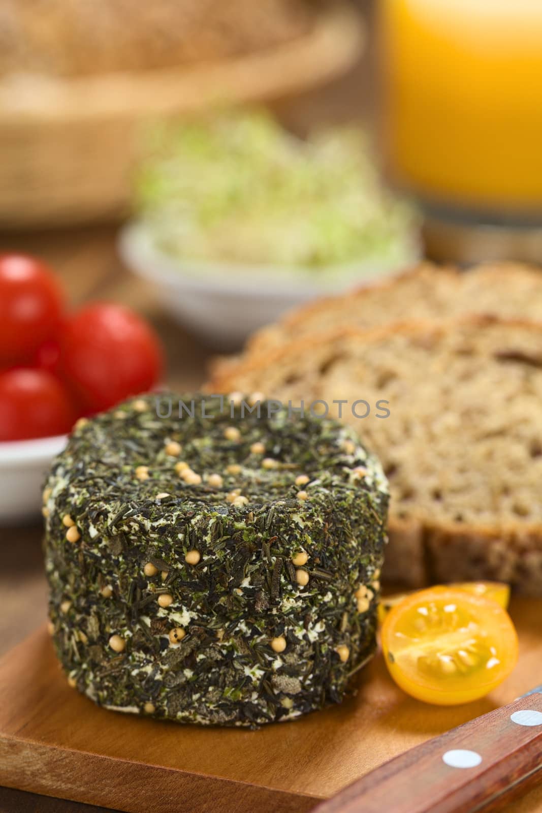 Goat cheese covered with herbs, yellow cherry tomato and slices of wholegrain bread on wooden board (Selective Focus, Focus on the upper edge of the cheese)