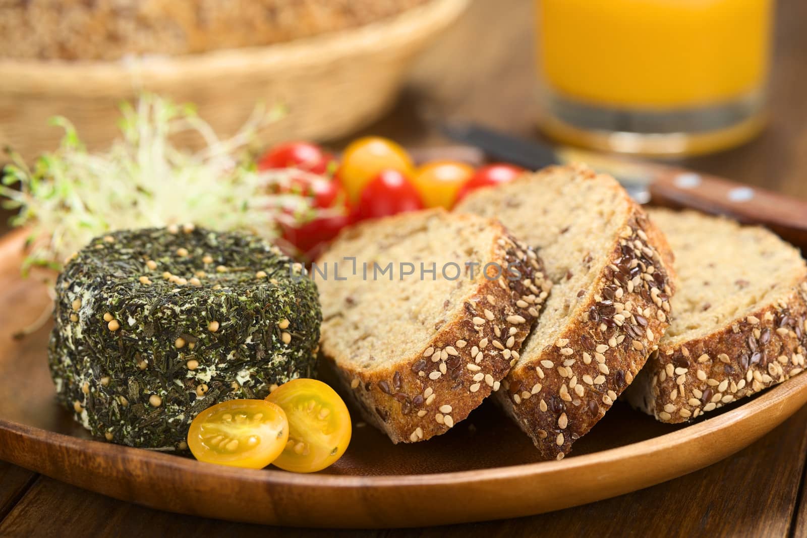 Goat cheese covered with herbs, yellow cherry tomato and slices of wholegrain bread on wooden plate (Selective Focus, Focus on the front of the cheese and the bread slices)