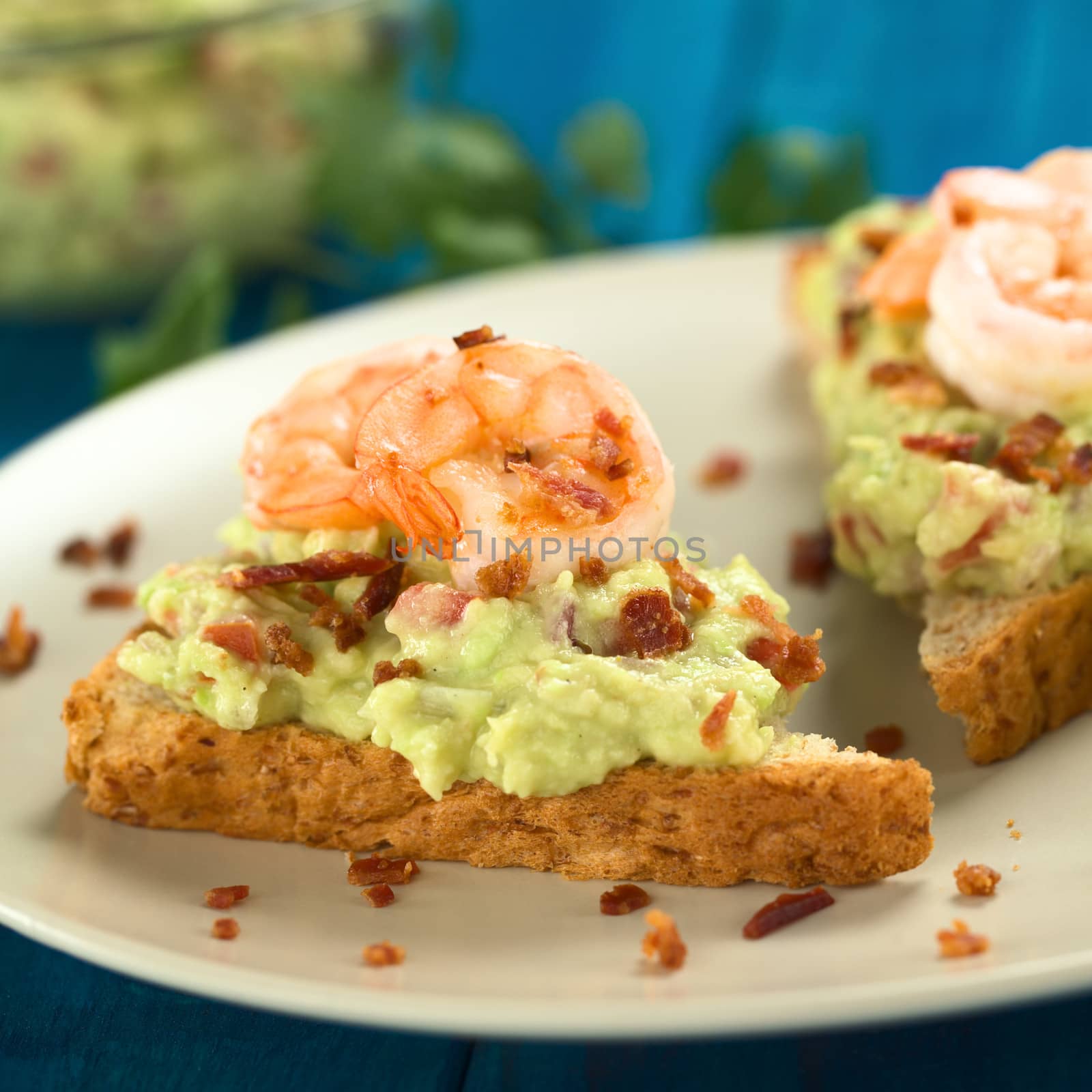 Wholegrain toast bread slices with guacamole, fried shrimp and fried bacon pieces (Selective Focus, Focus on the front of the shrimp on the bread) 