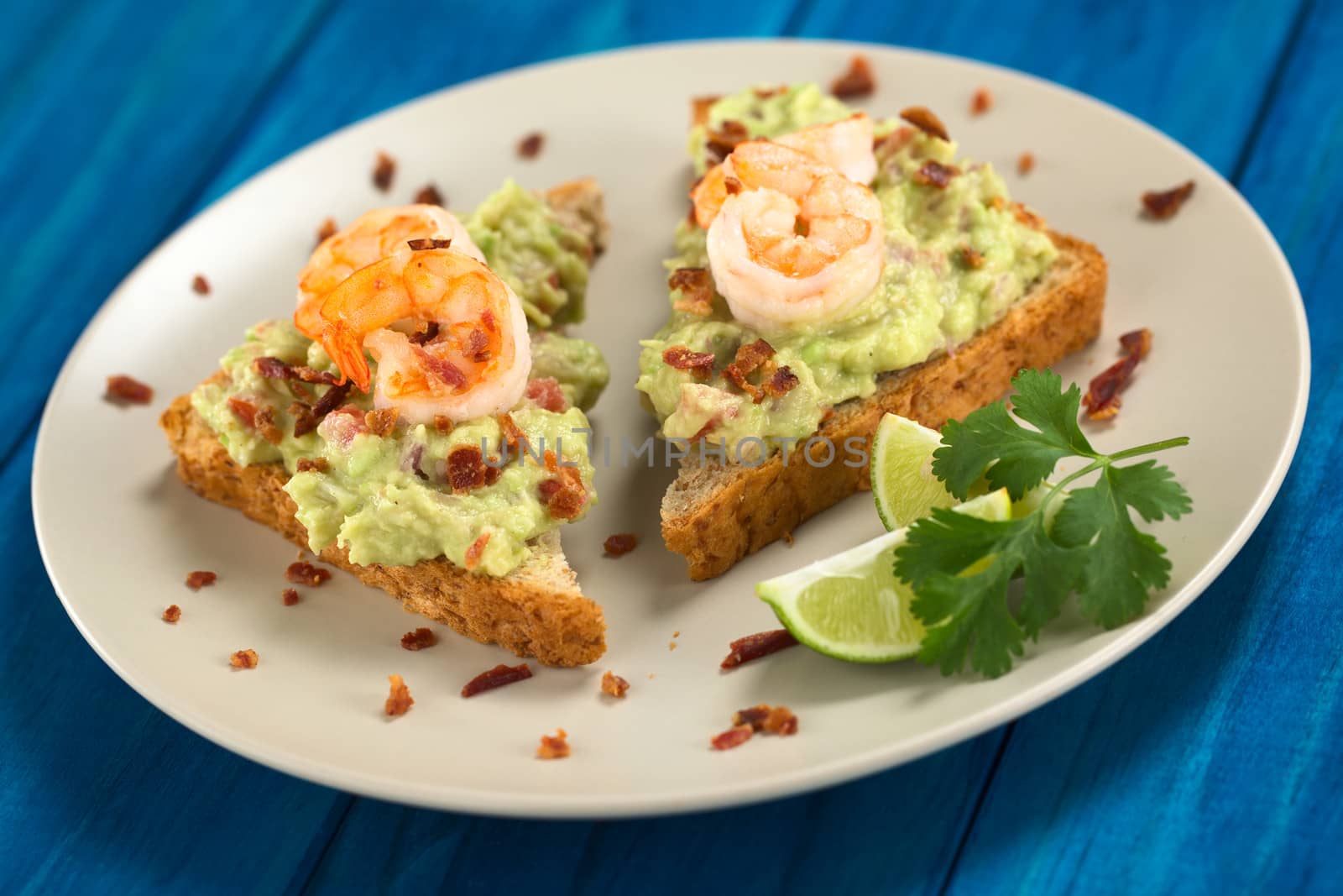 Wholegrain toast bread slices with guacamole, fried shrimp and fried bacon pieces served on plate on blue wood (Selective Focus, Focus on the front of the shrimp on the left bread and the front of the right bread) 