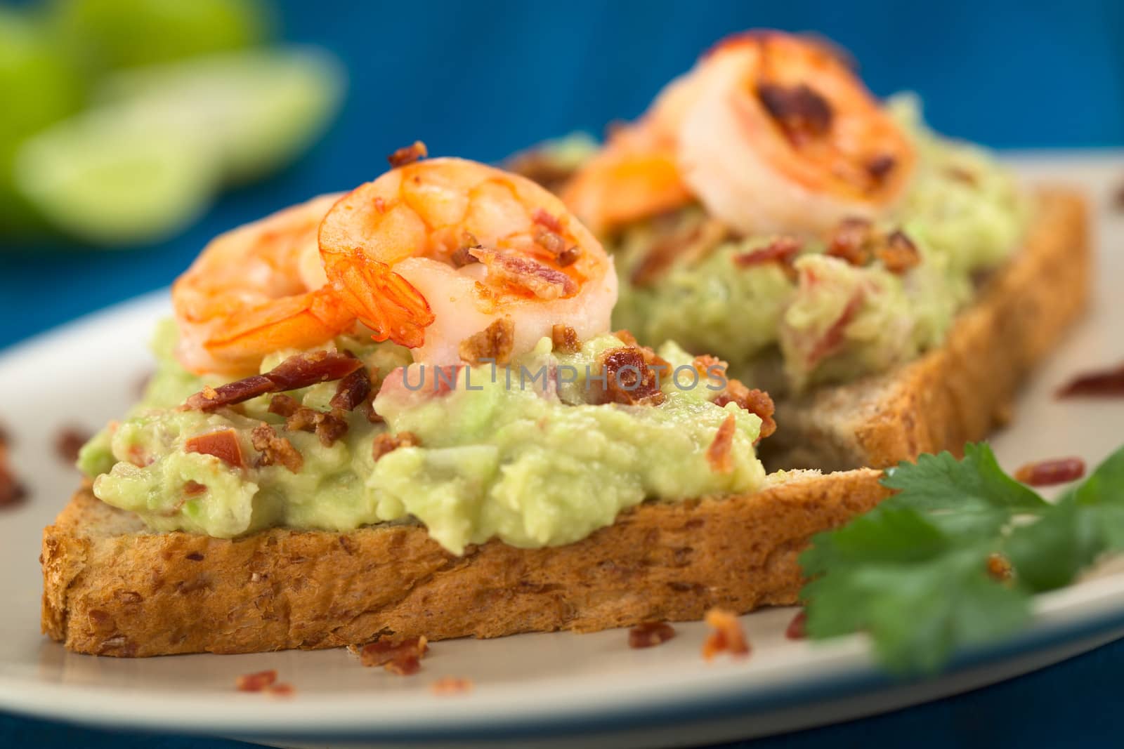 Wholegrain toast bread slices with guacamole, fried shrimp and fried bacon pieces served on plate on blue wood (Selective Focus, Focus on the front of the shrimp in the middle) 