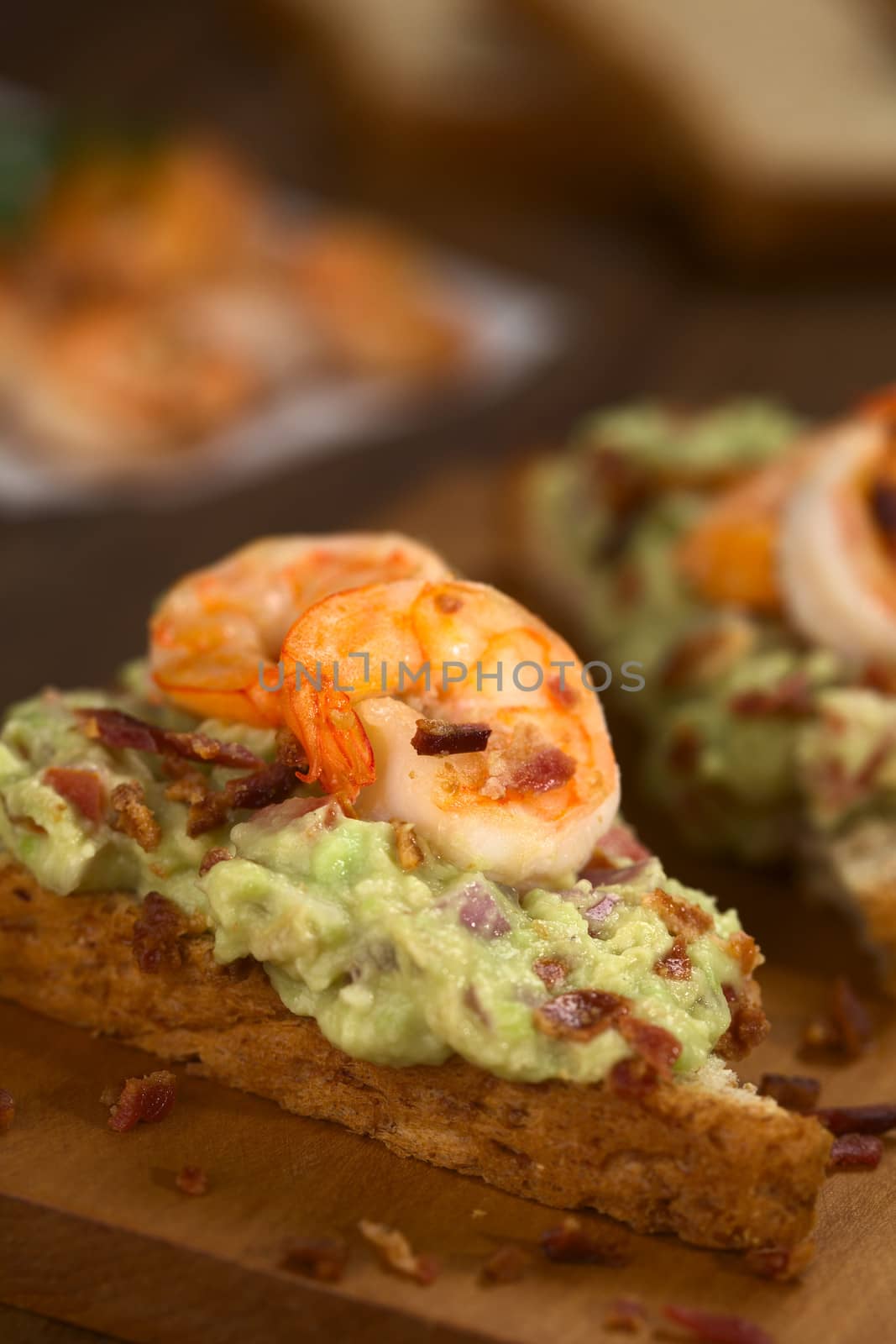 Wholegrain toast bread slices with guacamole, fried shrimp and fried bacon pieces on wooden board (Selective Focus, Focus on the front of the shrimp on the bread) 