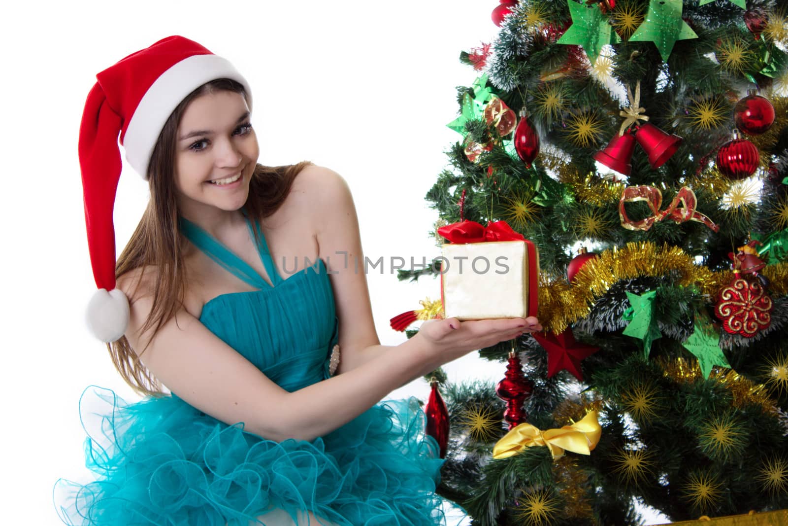 Smiling teenage girl in Santa hat offering present under Christmas tree over white