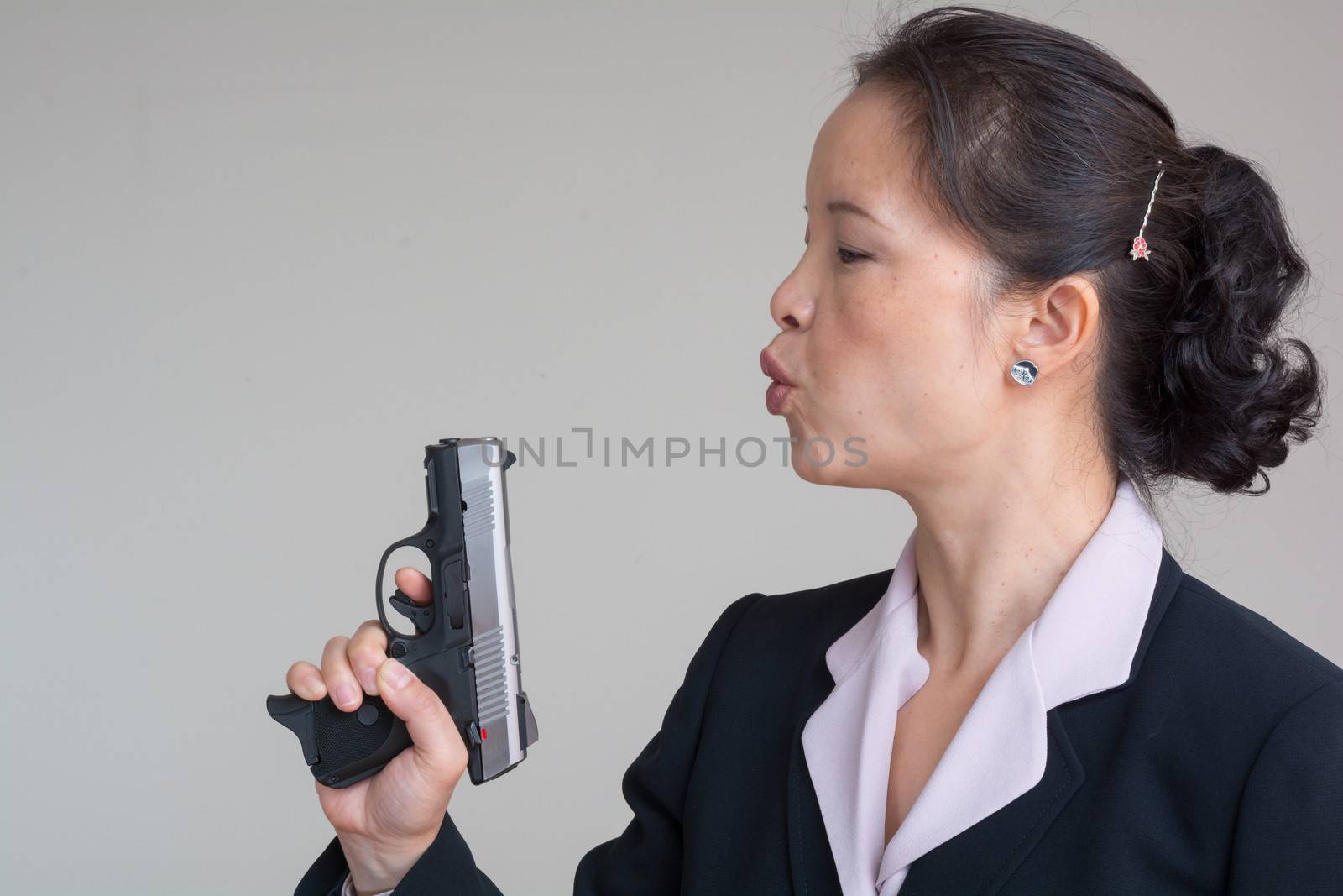 Woman in business suit blowing smoke off a fired hand gun on grey background