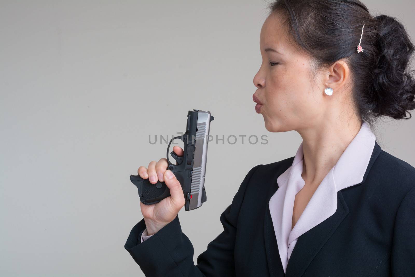 Woman in business suit blowing smoke off a fired hand gun on grey background