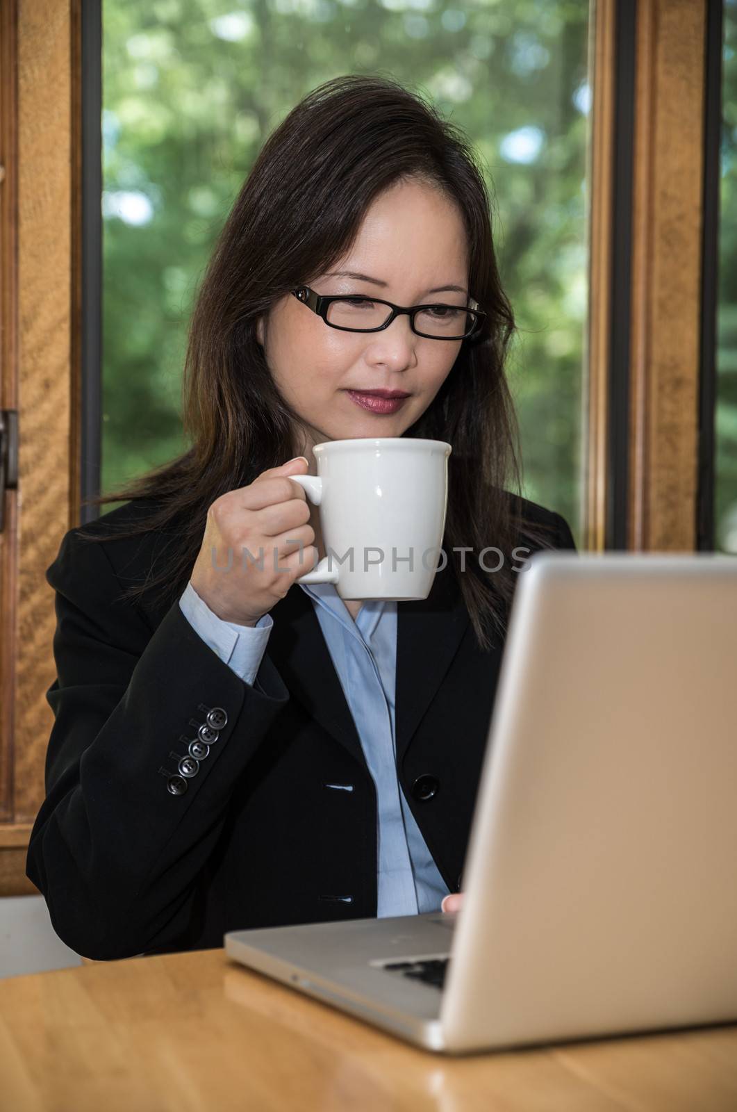 Woman in business suit with laptop on desk and drinking coffee in front of a window