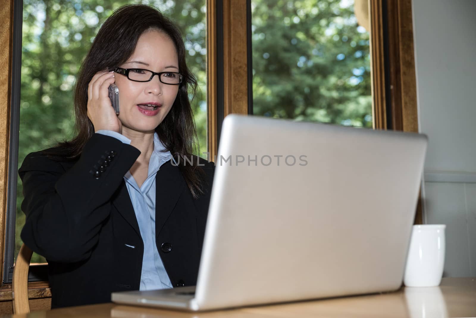 Woman in business suit with laptop and coffee on desk making a phone call in front of a window