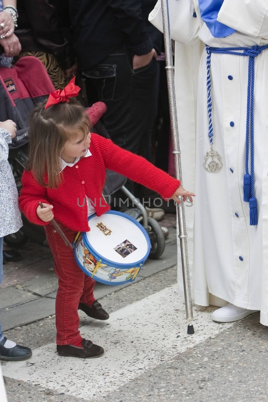 Penitent with staff of silver during a procession of holy week on Palm Sunday, Girl dressed in red with a toy drum looking in front of a Nazarene, Andalusia, Spain