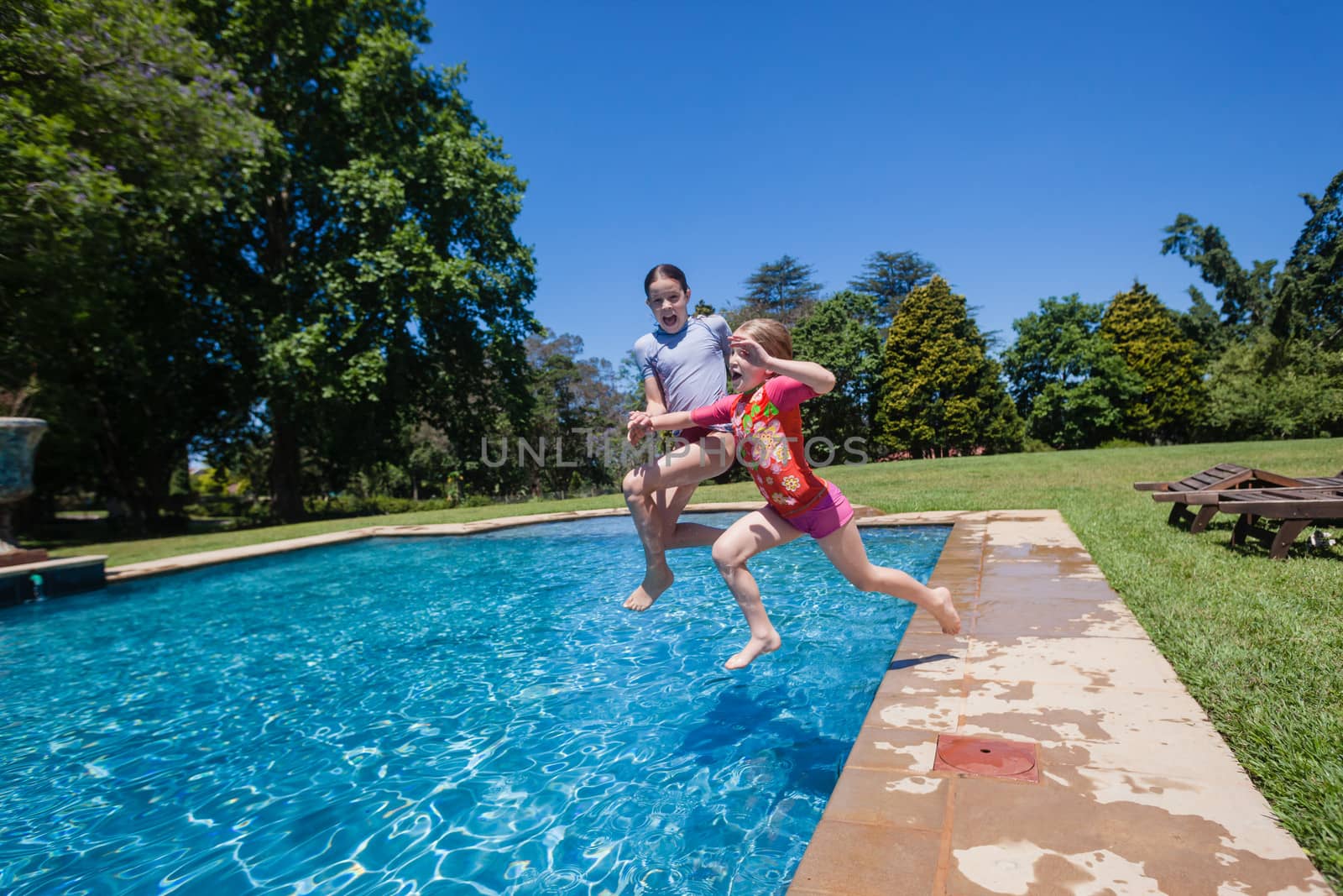 Young girls sisters having fun jumping into summer blue swimming pool.