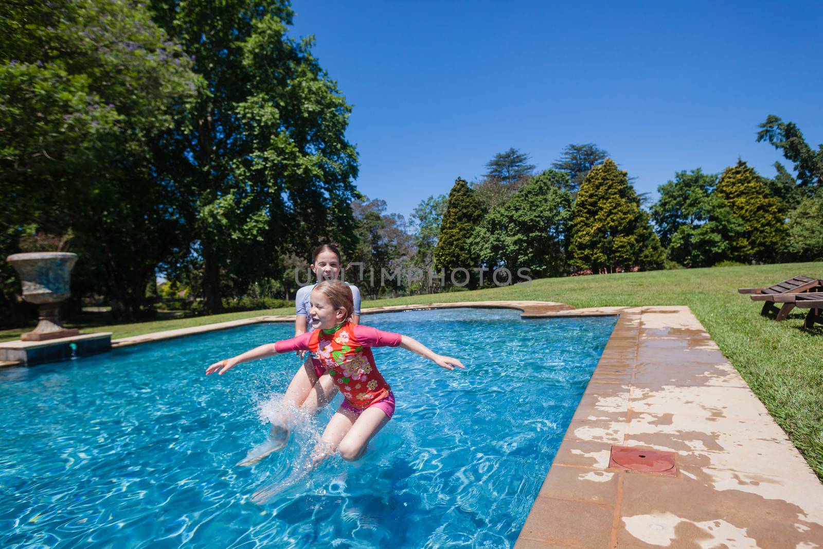 Young girls sisters having fun jumping into summer blue swimming pool