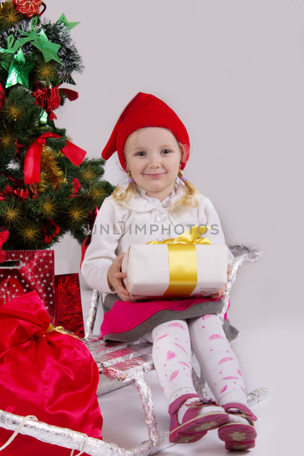 Little girl in Santa hat with present on sledge under Christmas tree