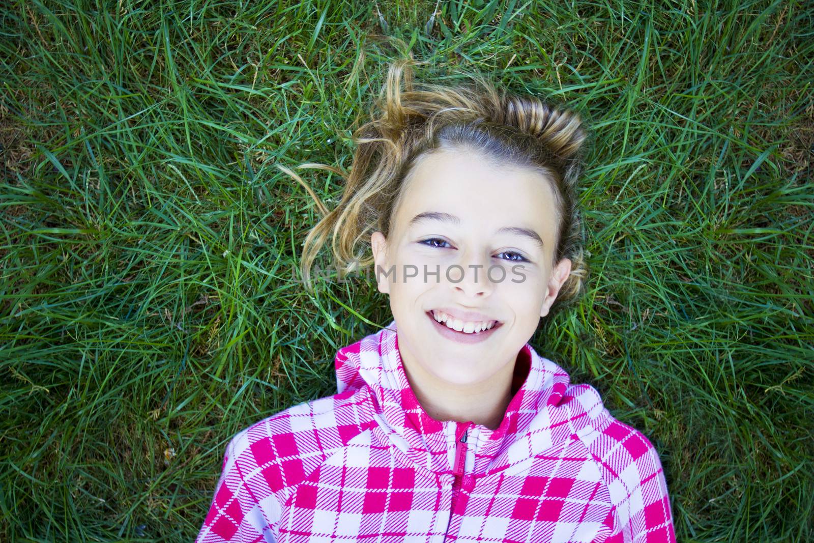 Teenage woman with checked pind white hoodie lying back on green grass, laughing and looking at the camera. Candid shot, real people