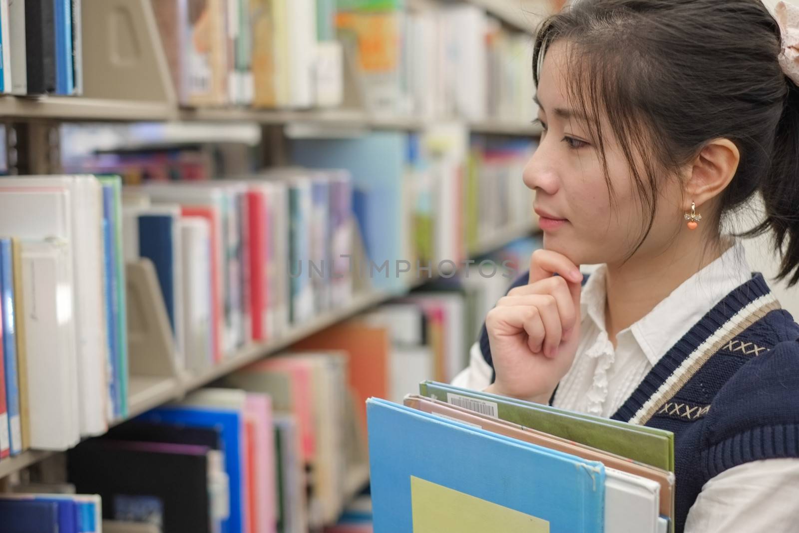 Cute young woman holding books and looking at a library bookshelf thoughtfully