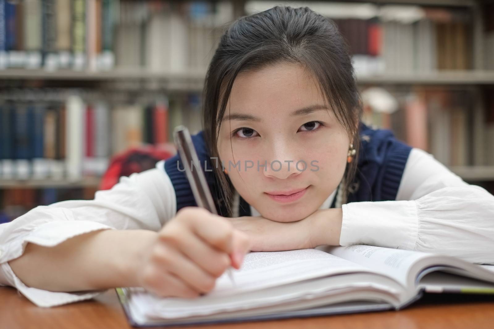 Tired young student resting her head on a textbook and doing homework in front of a library bookshelf