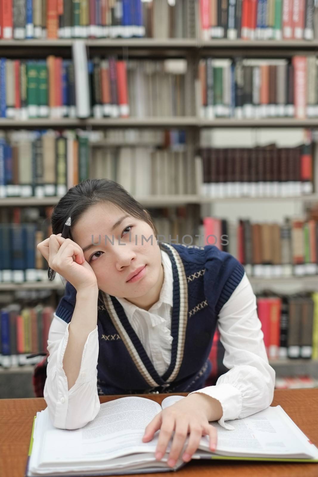 Young girl student with open textbook in front of a library bookshelf looking deep in thought