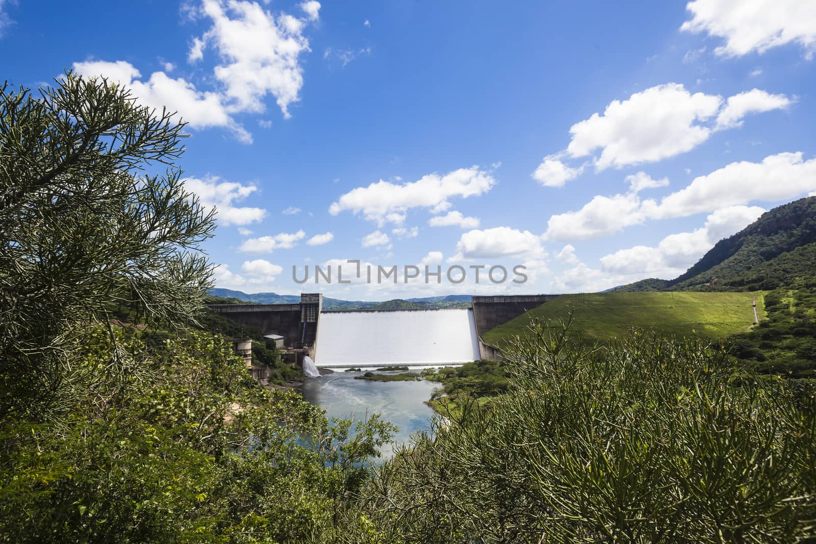 Dam filled to capacity and water flowing over the high wall in green summer valley.