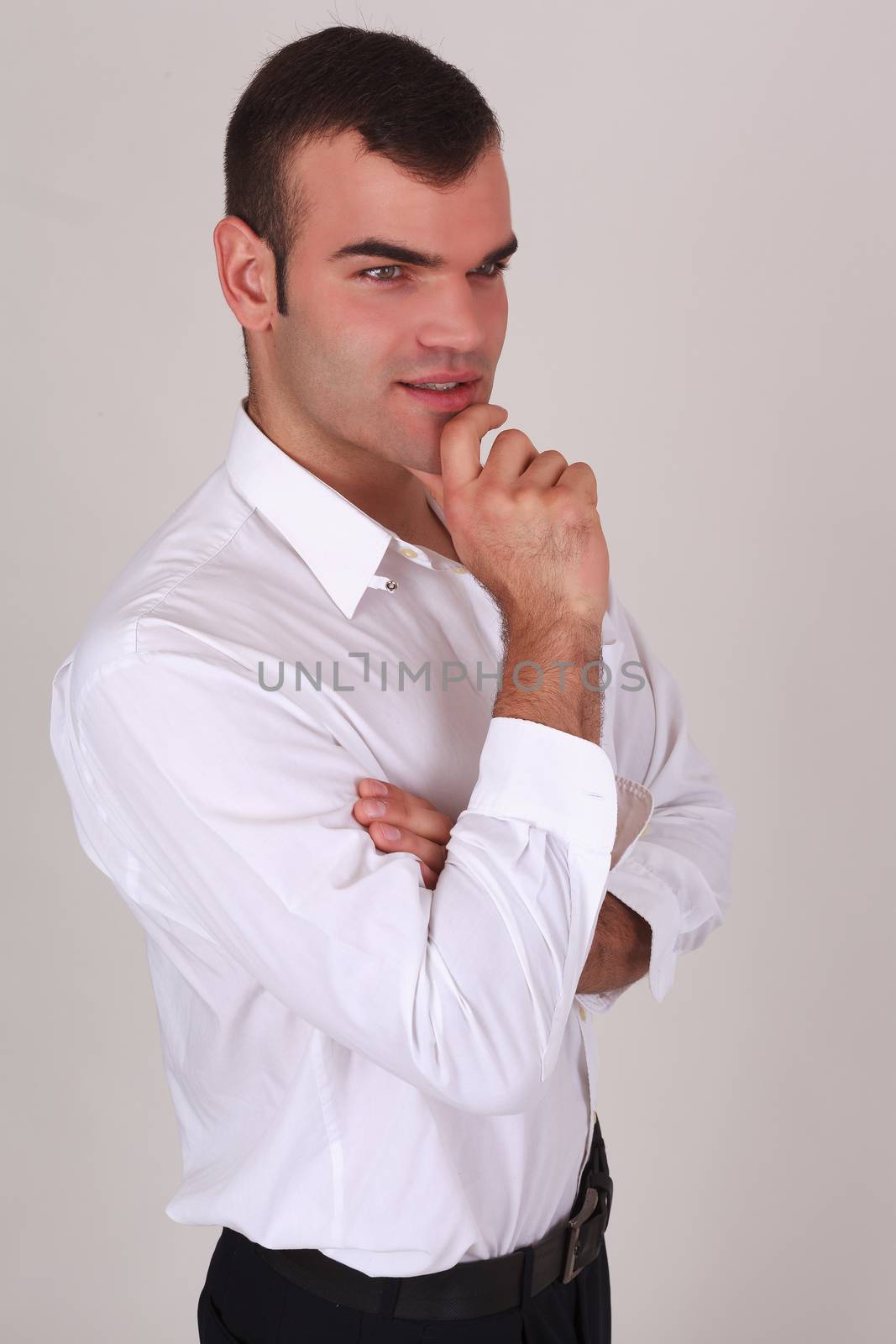 Handsome thoughtful, stylish man in a white shirt, smiling, with his arms crossed over his chest and his finger on the lip, side view on a gray background in studio
