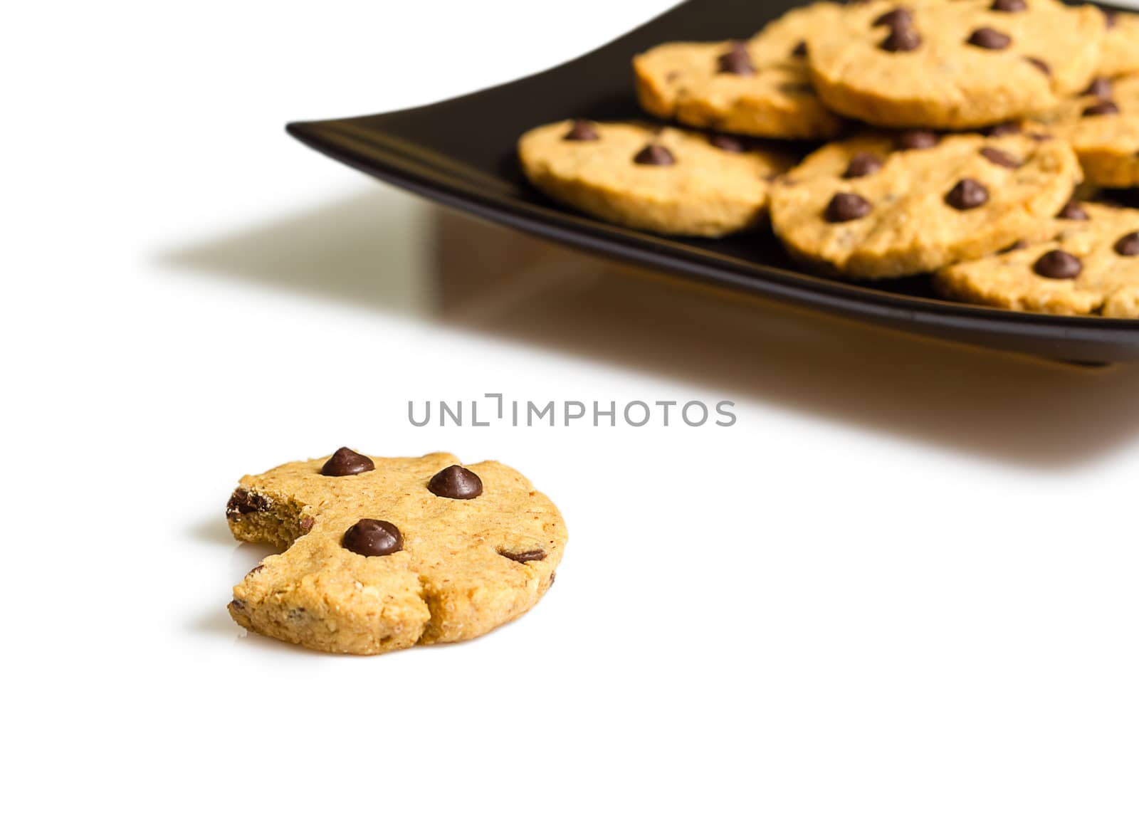 Closeup of chocolate chip cookie with a bite and pile of cookies in a square black plate on white background