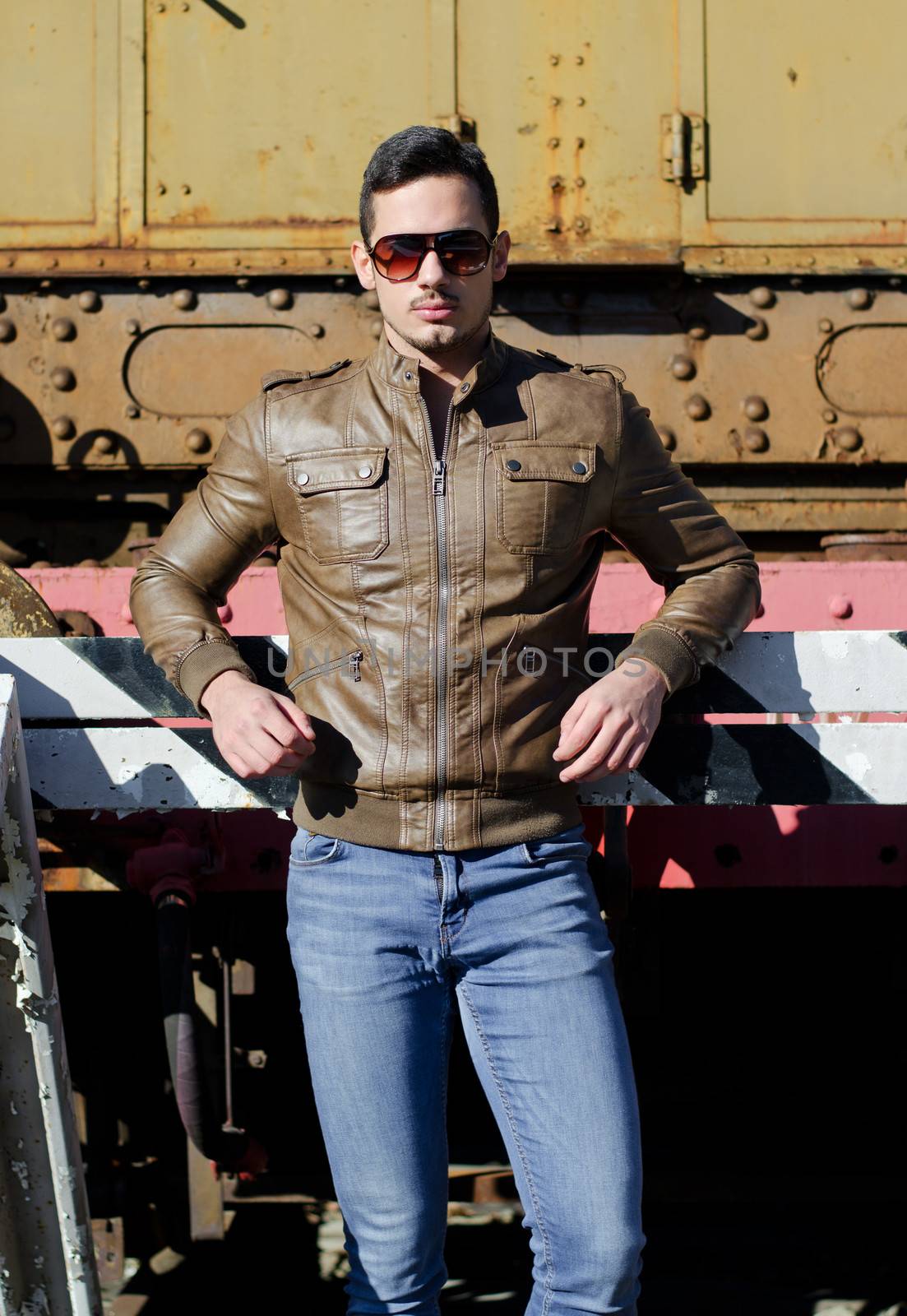 Attractive young man in leather jacket and jeans in front of old train wearing sunglasses