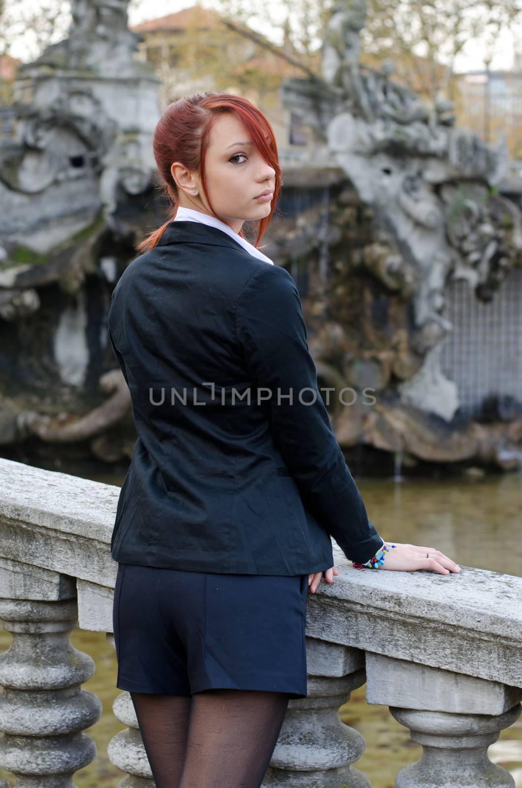 Elegant red-head girl outdoors against stone banister looking at camera over the shoulder, shot from the back